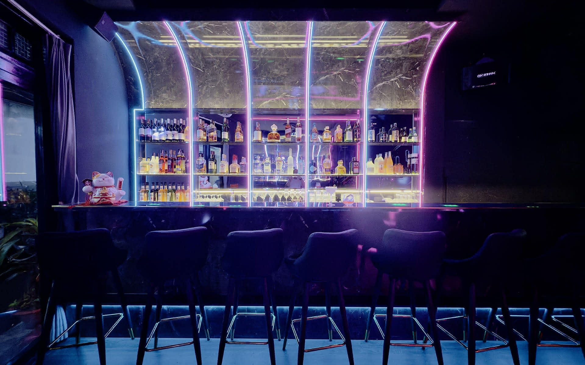 The bar at Barbie Bar: stools under lit arches and a backlit whisky wall in magenta and cyan neon.