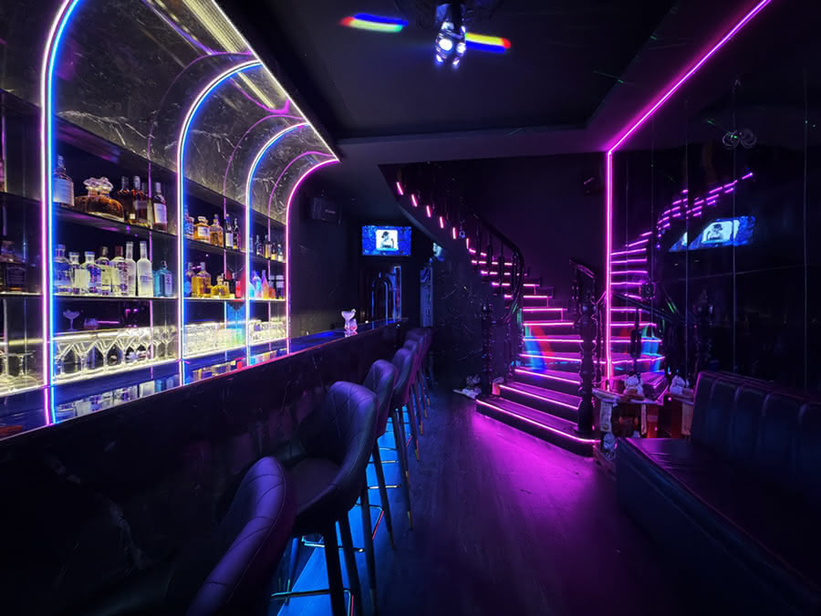 Inside Barbie Bar: a long bar counter, backlit bottle shelves, and a neon-edged staircase to the VIP rooms.